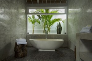 A modern bathroom with a freestanding bathtub, a window with plants, and a wooden stool with towels.