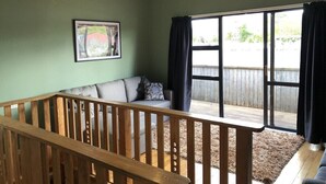 A room with a wooden railing, a sofa, a large window, and a view of a backyard. - The heart of Ohakune township with mountain views (Ohakune)