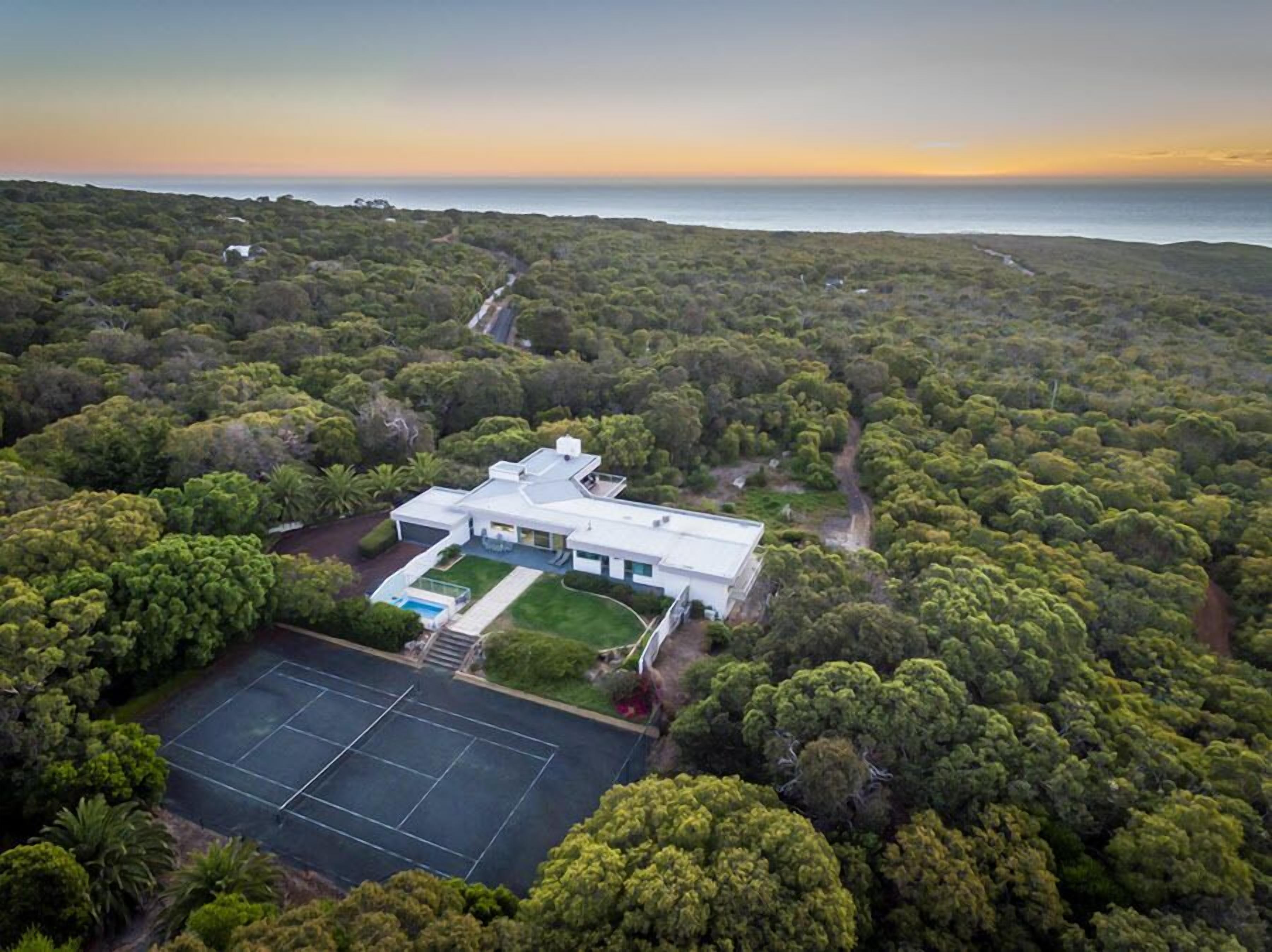 Vista aérea de uma casa moderna rodeada por uma floresta densa, com quadra de tênis e um caminho que leva ao oceano.