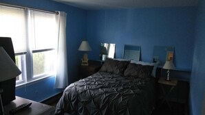 Un dormitorio con cama, dos mesitas de noche con lámparas, una ventana con persianas y un ventilador de techo.