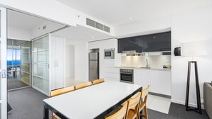 A modern kitchen with white cabinets, a stainless steel refrigerator, and a microwave. There is a dining area with a white table and wooden chairs. - Apartment 11504 is a holiday makers dream. Boasting a prime location (Surfers Paradise)