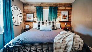 Ein Schlafzimmer mit Holzwandverkleidung, einer großen Wanduhr, einem Bett mit Metallgestell und einem Fenster mit Vorhängen.