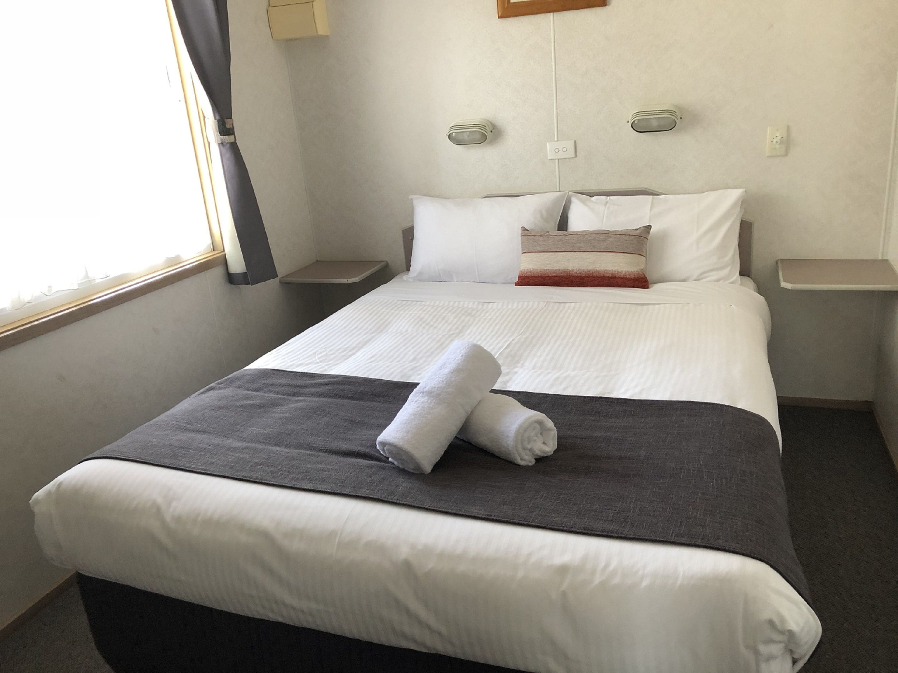 Een net opgemaakt bed met wit en grijs beddengoed, een opgevouwen handdoek en een nachtkastje met een lamp.