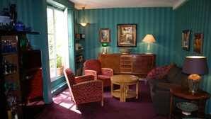 Ein Zimmer mit türkisfarbenen Wänden, einem Holzschrank, einem rot gemusterten Sessel, einem runden Holztisch und einem violetten Sofa.