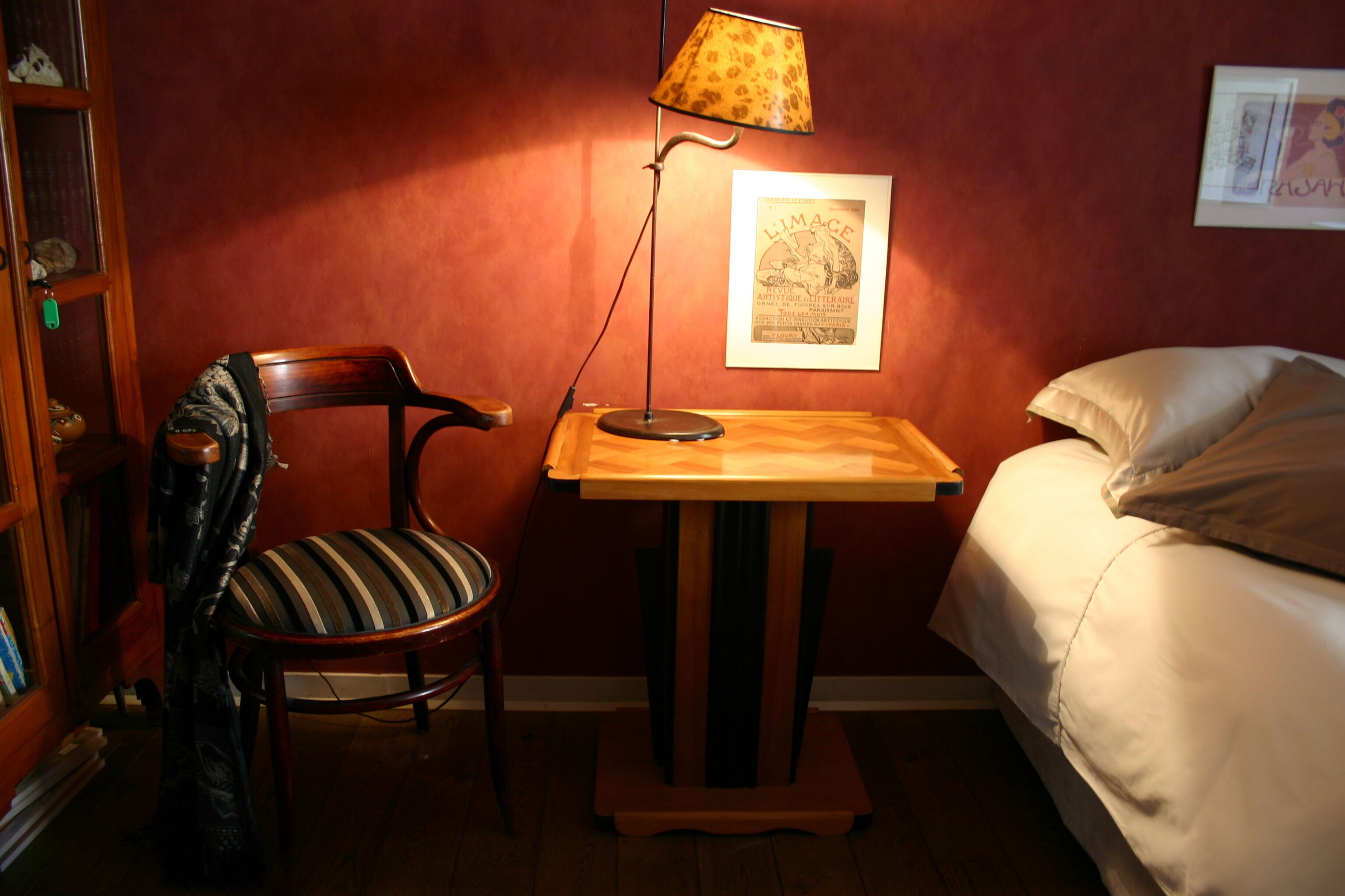 Een kamer met een houten stoel, een klein tafeltje met een lamp, een bed en een lijst met een foto aan de muur.