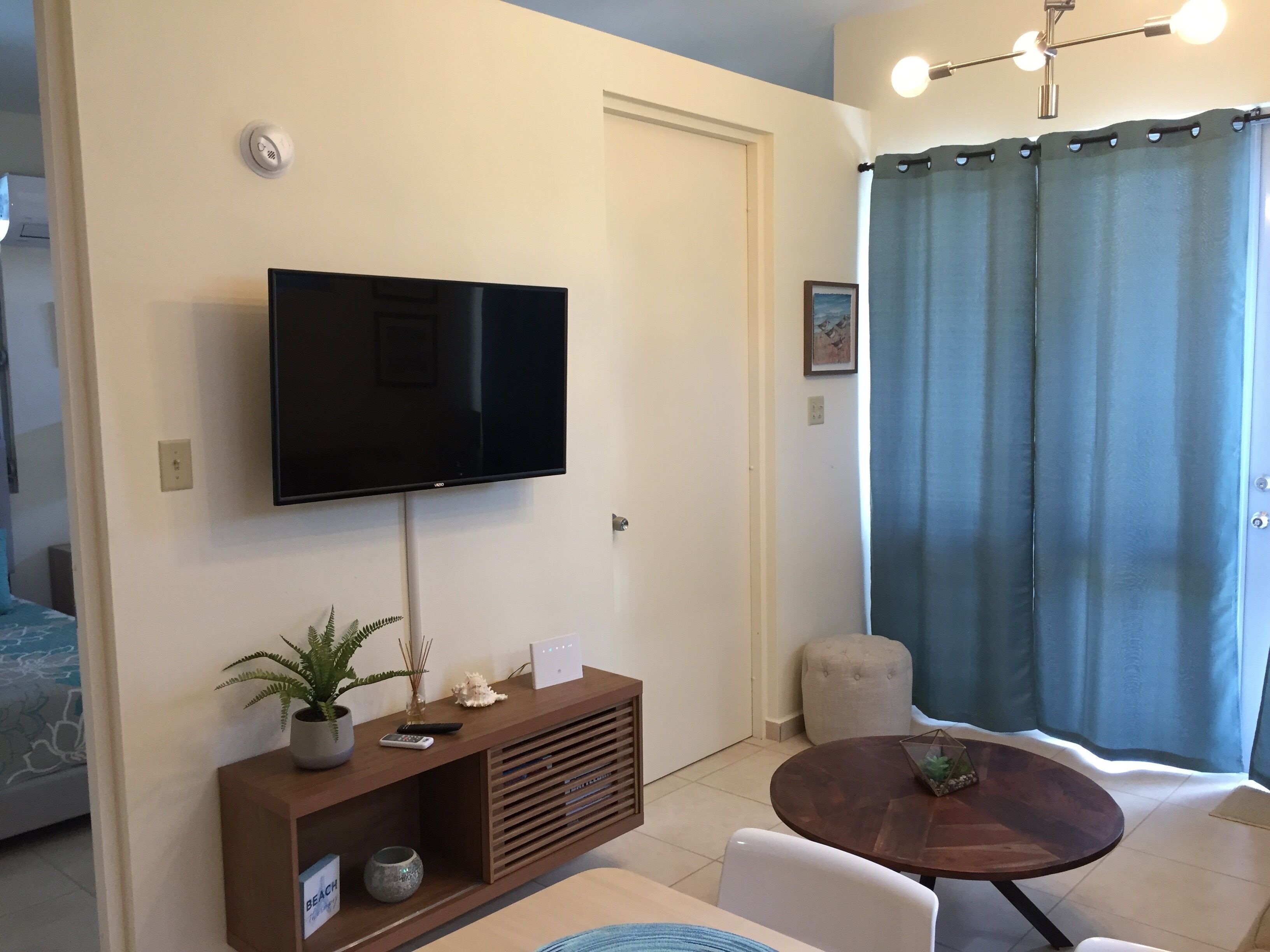 Una sala moderna con televisor de pantalla plana montado en la pared, un mueble de madera para entretenimiento, una mesa redonda de centro y una cortina azul.