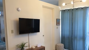 Una sala de estar con un televisor de pantalla plana montado en la pared, una pequeña consola de madera con una planta y un control remoto, una mesa redonda de centro, y una cortina azul cubriendo una ventana.