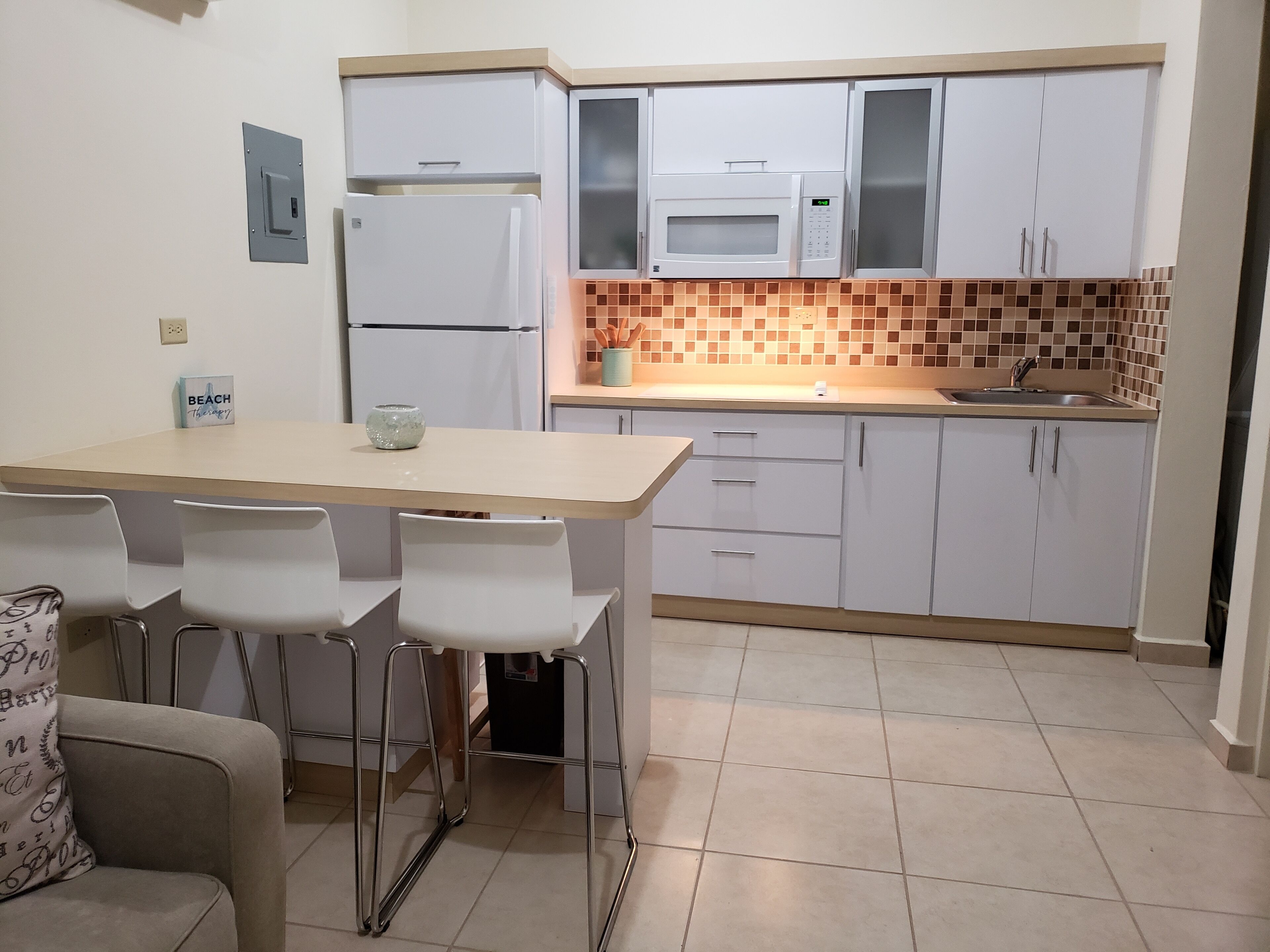 Une cuisine compacte avec des meubles blancs, un four à micro-ondes, un réfrigérateur et un plan de travail recouvert de carreaux. On y trouve également un petit coin repas avec une table et des chaises.