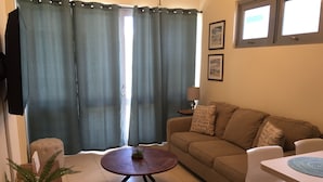 Una sala de estar con un sofá, una mesa de centro, una mesa de comedor, una planta en maceta y una ventana con cortinas.