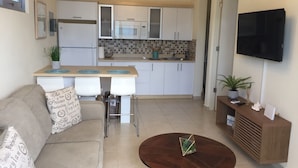 Una cocina moderna con armarios blancos, un microondas y un lavabo. Hay una mesa de comedor con sillas, un sofá con cojines estampados y una mesa de centro de madera con un objeto decorativo. Un televisor está montado en la pared y hay una planta sobre una mesita auxiliar.