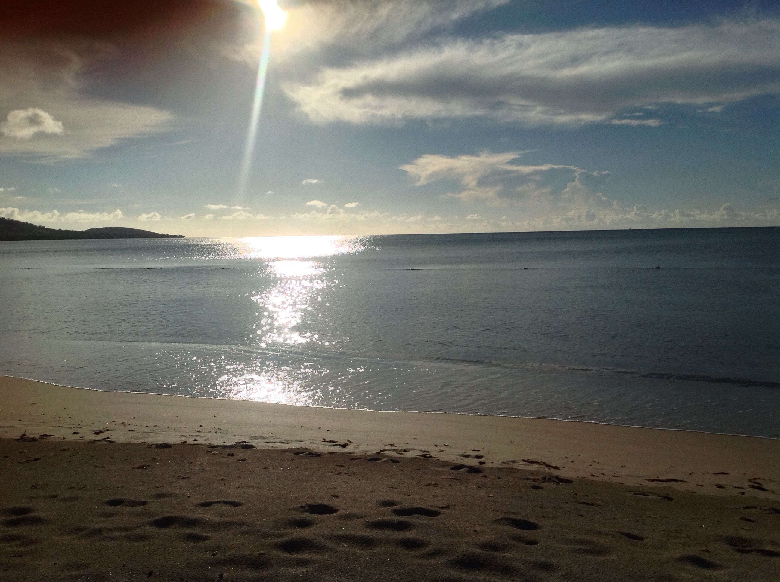 A beach with footprints leading to the water, a clear sky, and the sun reflecting on the surface.