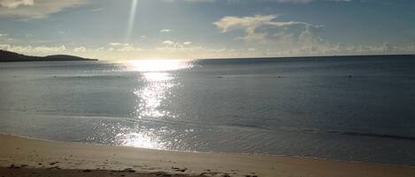 A beach with footprints leading to the water, a clear sky, and the sun reflecting on the surface.