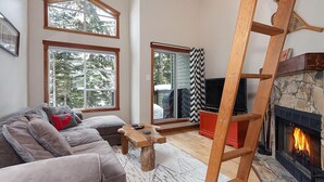 Tunnelmallinen olohuone, jossa on takka, puinen tikapuu, sohva ja suuret ikkunat.