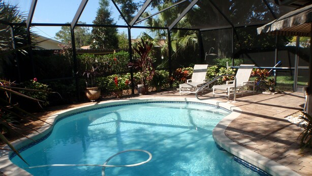 A clear swimming pool with a tiled edge, surrounded by a covered patio area with lounge chairs and potted plants.