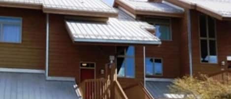 A multi-story wooden building with a metal roof, a staircase leading to the entrance, and a small porch area.