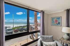 A room with a large window overlooking a beach, a chair, a lamp, and a painting on the wall.