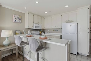 Une cuisine moderne avec des meubles blancs, un îlot central doté de tabourets, un four à micro-ondes, un réfrigérateur et une table à manger avec une lampe.