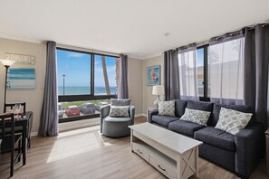 A modern living room with a sofa, armchair, and dining table, large windows with curtains, and a view of the sea.