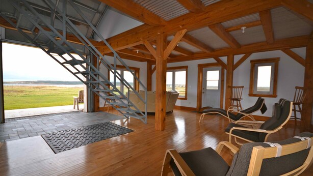 A spacious room with a wooden floor, a staircase leading to a balcony, and several reclining chairs. - New  Beachfront  Post+beam Cottage, Amazing Sunsets and Just Off Cabot Trail (Point Cross)