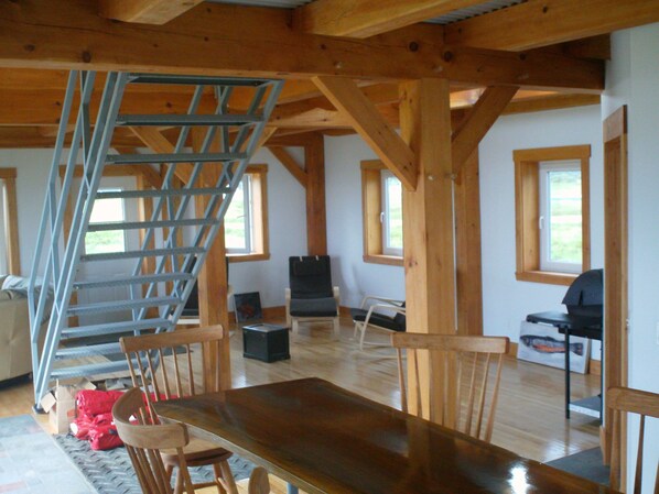 A spacious room with a wooden dining table, chairs, a staircase, and a black couch. - New Beachfront Post+beam Cottage, Amazing Sunsets and Just Off Cabot Trail (Point Cross)