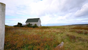 A white house with a green roof, surrounded by a grassy field with some bushes and a wooden fence. - New  Beachfront  Post+beam Cottage, Amazing Sunsets and Just Off Cabot Trail (Point Cross)