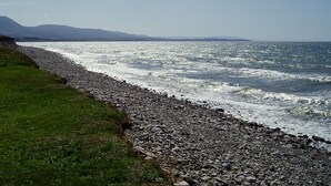 A rocky shoreline with waves crashing onto the beach, a grassy area, and distant mountains under a clear sky. - New  Beachfront  Post+beam Cottage, Amazing Sunsets and Just Off Cabot Trail (Point Cross)