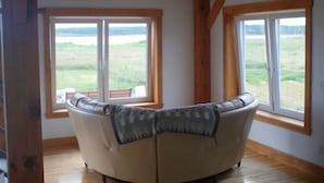 A room with a curved sofa, two large windows with a view of a grassy field, wooden beams on the ceiling, and a wooden floor. - New  Beachfront  Post+beam Cottage, Amazing Sunsets and Just Off Cabot Trail (Point Cross)