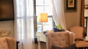 A living room with two beige armchairs, a glass coffee table, a white desk with a lamp, and sheer curtains. - Location, Location, Location!  Carriage House in Historic Old NE District (St. Petersburg)