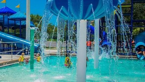 A water play area with multiple water jets and slides, surrounded by a fenced area with umbrellas and a playground in the background. - Location, Location, Location!  Carriage House in Historic Old NE District (St. Petersburg)
