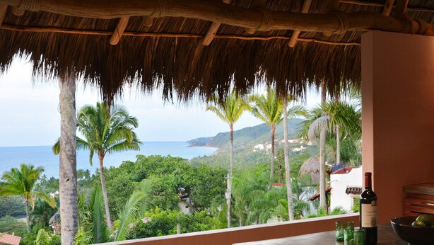 A room with a view of the ocean and palm trees, a table with a bottle and glasses, and a thatched roof. - Casita Mimas: High design, great views, romantic retreat  (Sayulita)