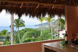 A room with a view of the ocean and palm trees, a wooden table, and a bottle of wine.