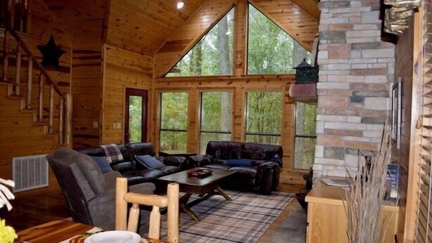 Interior acogedor de una cabaña con techo de madera, chimenea de piedra, ventanas amplias y una zona de estar confortable.