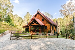 A rustic wooden cabin with a porch, surrounded by a gravel driveway and a wooden fence.