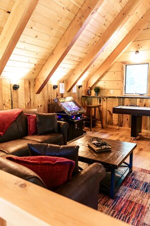 A cozy room with a wooden ceiling, a pool table, a sofa with cushions, and a gaming console.