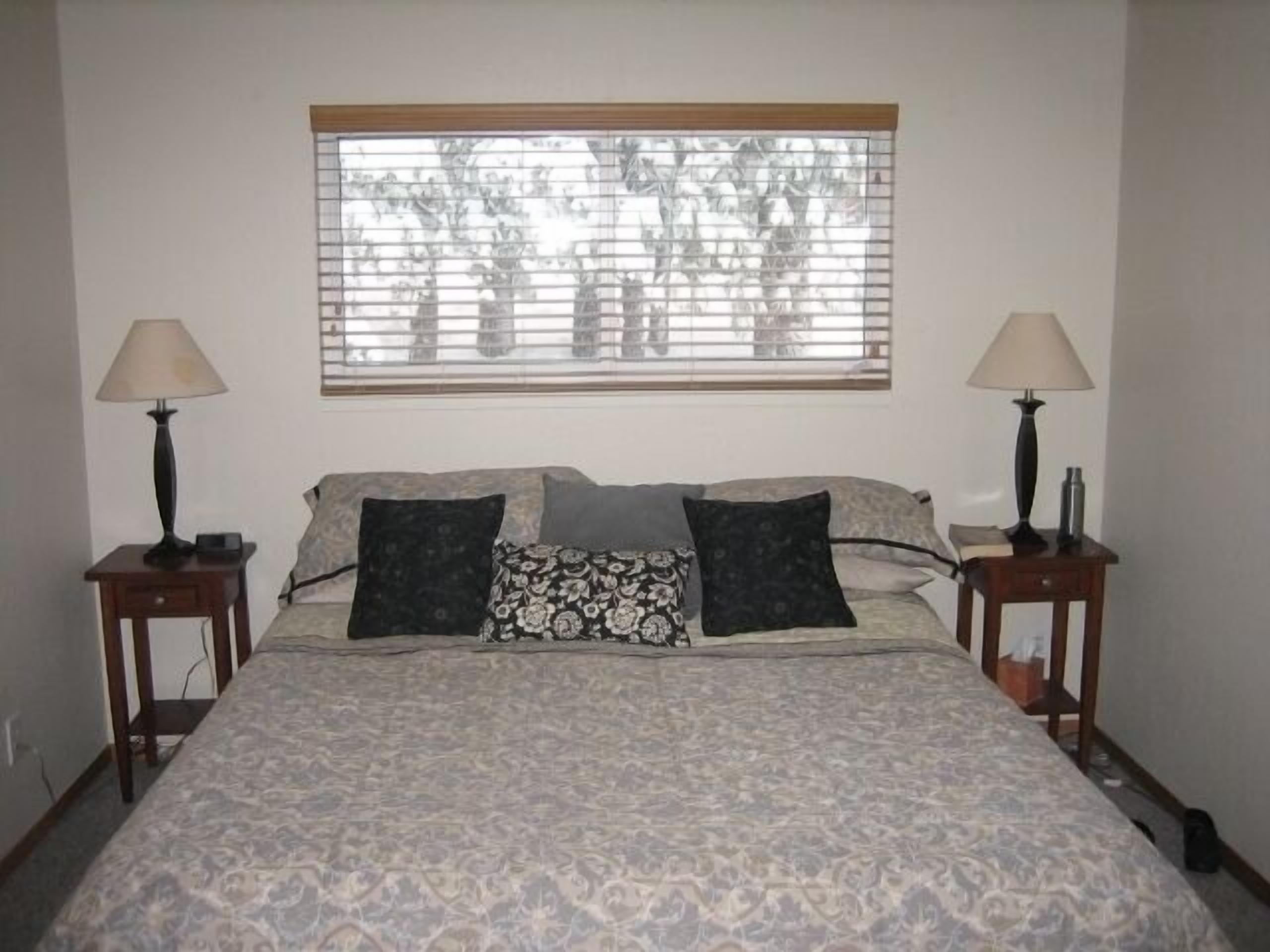 A bedroom with a large bed, two bedside tables with lamps, a window with blinds, and a patterned bedspread.