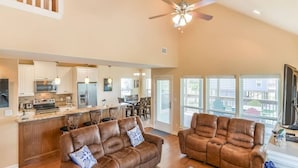 Amplia sala de estar con un sofá y un sillón a juego, ambos de cuero marrón, una cocina con muebles blancos y un ventanal con vistas al exterior.