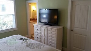 A bedroom with a bed, a dresser with drawers, a television on top, a door, and a window with blinds. - Bay Point Experience (Marblehead)