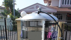 Ein großer, metallener Wassertank mit Kuppeldeckel, verbunden mit einem kleineren Tank und einer Steuereinheit, befindet sich auf einem Balkon mit Metallgeländer und Fliesenboden.