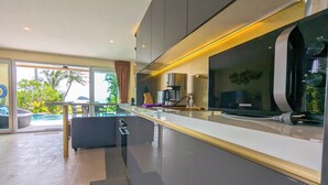 Een moderne keuken met een groot flatscreen televisietoestel, een koffieapparaat en uitzicht op een zwembad en groen.