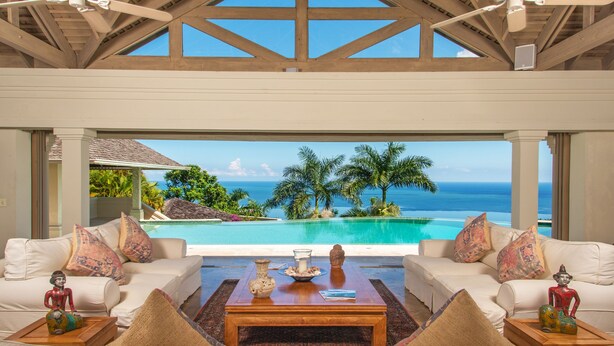 A luxurious room with a large window overlooking a pool and the ocean. There is a white sofa with colorful cushions and a wooden coffee table in the center. - Silent Waters Villa - 9BR/7BTH estate - ocean and river views - fully staffed (Montego Bay)