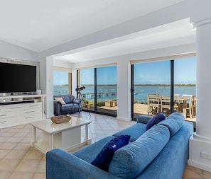 En moderne stue med stort fjernsyn, sofa og sofabord. Der er en balkon med havudsigt.