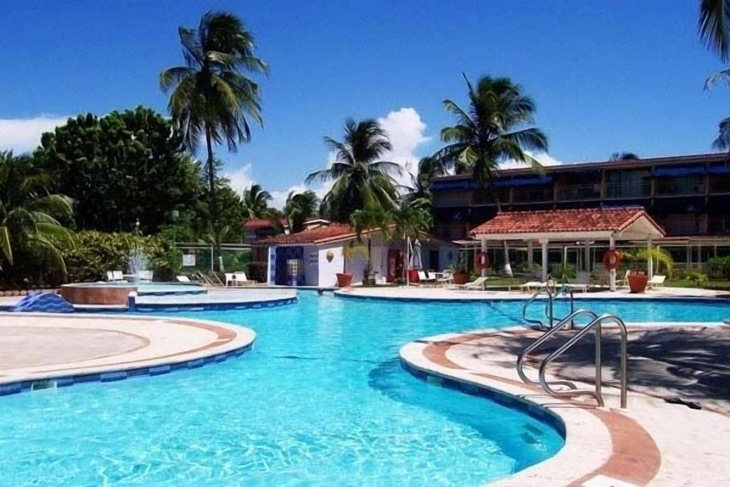 Una piscina con varias pistas, palmeras y un edificio de techo rojo al fondo.