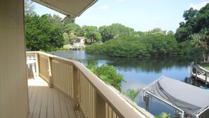 A wooden deck with a railing overlooking a calm river, surrounded by lush greenery and a dock with a covered area.