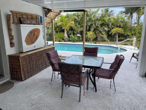 A covered outdoor seating area with a glass table and chairs, a tiki bar sign, and a pool with palm trees in the background.