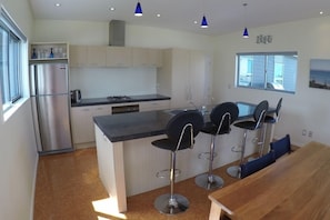 A modern kitchen with a central island, bar stools, and a wooden dining table. - Relax in Rangiputa - Decks, Beaches and Boating (Rangiputa Beach)