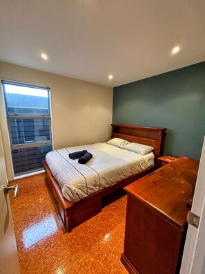 A bedroom with a bed, a wooden nightstand, a window, and a door. - Relax in Rangiputa - Decks, Beaches and Boating (Rangiputa Beach)