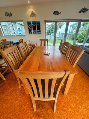 A dining area with a wooden table and chairs, a decorative wall piece, and a view of the outdoors through large windows. - Relax in Rangiputa - Decks, Beaches and Boating (Rangiputa Beach)