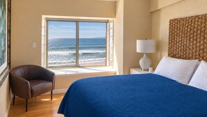 Ein Hotelzimmer mit großem Fenster und Meerblick, einem Bett mit blauer Bettdecke, einem braunen Ledersessel und einem gewobenen Kopfteil.