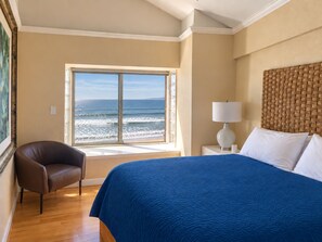 Una habitación de hotel con un ventanal que ofrece vistas al océano, una cama con colcha azul, un sillón de cuero marrón y un cabecero tejido.