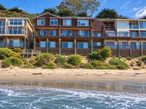 Un hotel frente a la playa con varios balcones, fachada de madera y una playa arenosa al frente.